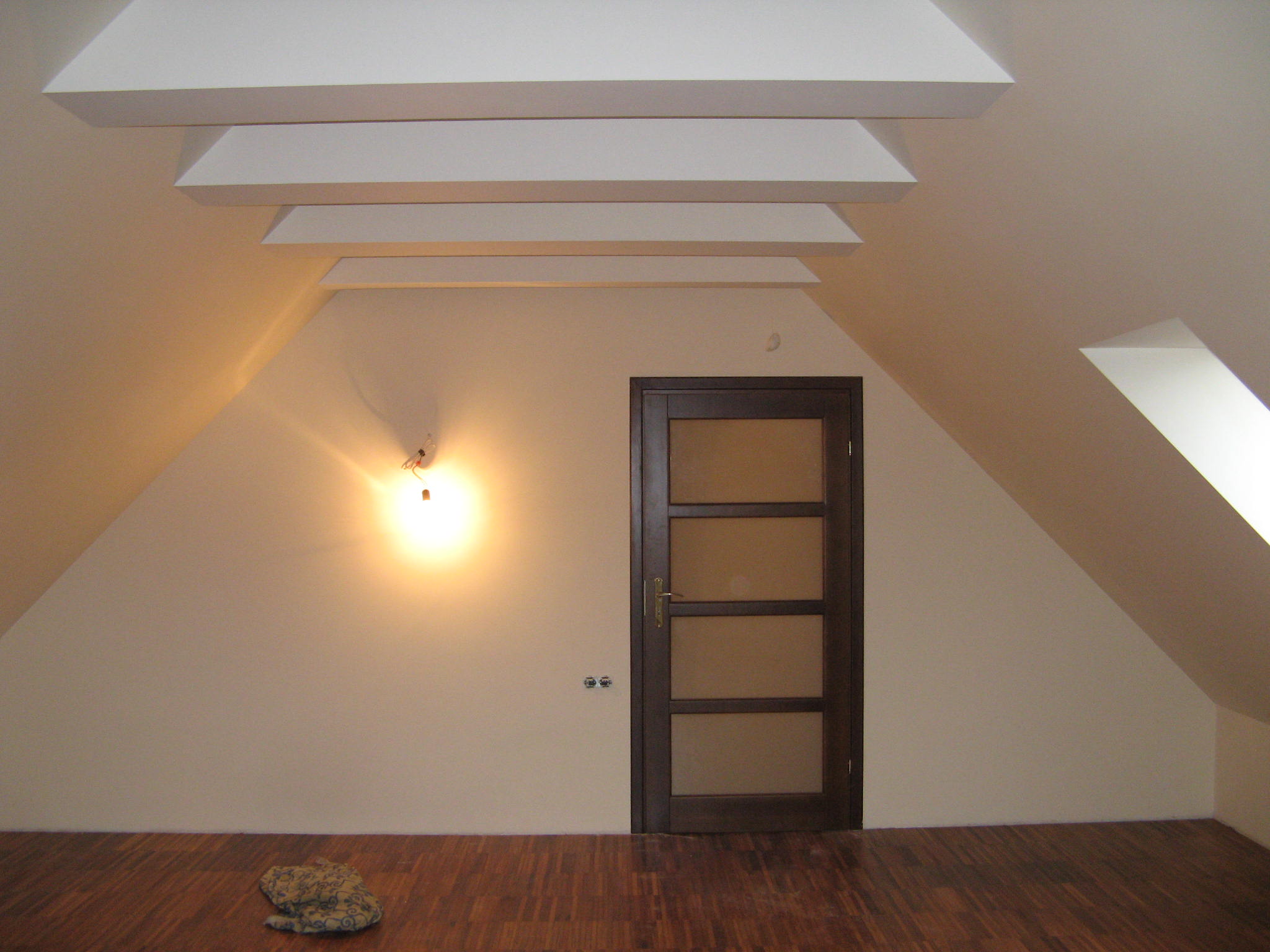 Poddasze z ukośnymi ścianami, drewnianą podłogą, brązowymi drzwiami z szybami i dekoracyjnymi, białymi belkami na suficie. Na ścianie widoczne gniazdka elektryczne i żarówka bez klosza. Na podłodze...